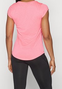 Kvinna som bär en figursydd rosa kortärmad skjorta och svarta träningsleggings, stående med ryggen mot kameran.