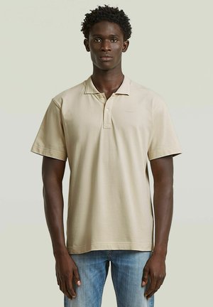 Man met kort krullend haar, gekleed in een beige polo met korte mouwen en een blauwe spijkerbroek, tegen een effen achtergrond.