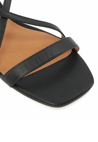 Sandale en cuir noir avec un design à bout carré, dotée de fines lanières croisées et d'une semelle intérieure beige. Texture lisse avec un minimum de détails.