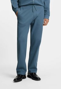 Blaue Nadelstreifenhose aus weichem Stoff, mit elastischem Bund und Kordelzug sowie einem lockeren Schnitt. Getragen mit schwarzen Schuhen.