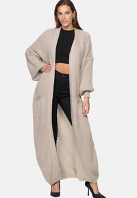 Long beige knit cardigan with open front, large balloon sleeves, two front pockets, and ribbed trim. Worn over a black cropped top and pants.