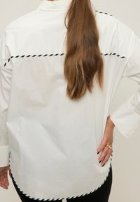 Weißes Hemd mit einem schwarz-weißen gedrehten Besatz entlang der Schultern und dem Saum, mit einer lockeren Passform und geknöpften Manschetten.
