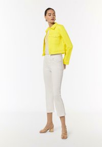 Yellow denim jacket with cropped design, two chest pockets, white t-shirt underneath, paired with white ankle-length jeans and beige heeled sandals.