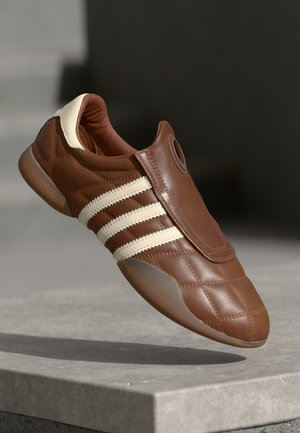 Baskets en cuir marron avec des rayures blanches et un col rembourré, présentées en suspension au-dessus d'une surface en pierre grise sur un fond sombre.