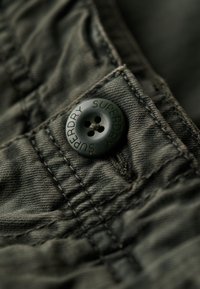 Tissu vert foncé avec une texture, présentant un grand bouton en plastique insculpé avec "SUPERDRY" et des détails de couture visibles.