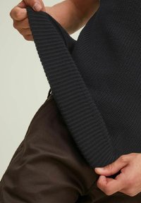 Dunkel gestrickter Pullover, der gehalten wird, um einen strukturierten, gerippten Saum zu zeigen. Er wird mit braunen, glatten Stoffhosen kombiniert.