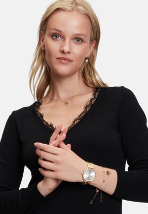 Schwarzer V-Ausschnitt-Pullover mit Spitze, geschichtete goldene Kettenhalsketten, herzförmige Ohrringe und eine goldene Uhr mit silbernem Ziffernblatt.