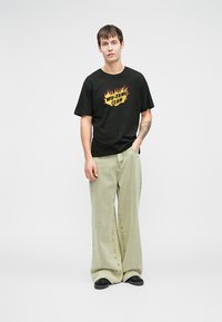 Μαύρο βαμβακερό T-shirt με "Wu-Tang Clan" σε κίτρινες φλόγες, συνδυασμένο με υπερμεγέθη ανοιχτό πράσινα τζιν παντελόνια και μαύρα παπούτσια.