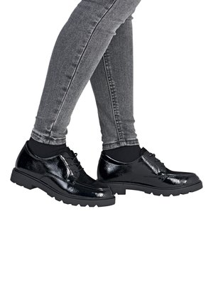 Legs wearing gray skinny jeans, black socks, and shiny black lace-up shoes with textured soles crossing on a white background.