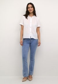 Hvid linned bluse med korte ærmer, rynket krave og knaplukning, parret med lyseblå tætsiddende jeans og beige hæle.