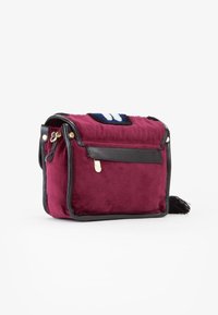 Piccola borsa a mano rettangolare in velluto bordeaux con rifiniture in pelle nera, zip dorata, applique decorativa e dettagli con frange su uno sfondo bianco.
