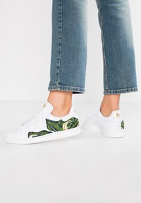 Valkoiset tennarit, joissa on vihreä lehtikuvio sivuilla, teksturoitu nahkapäällinen, pyöreä kärki ja kultaiset logokoristeet kannassa.