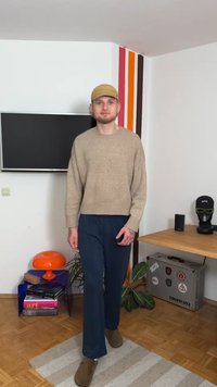 Beżowy sweter z dzianiny, luźny krój; granatowe szerokie spodnie; brązowe buty. W tle telewizor, pomarańczowa lampa i kolorowe paski na ścianie.