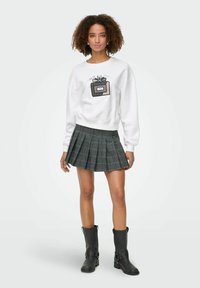 Bijela majica s kapuljačom s grafičkim dizajnom, uparena s naboranom sivo-kariranom suknjom i crnim gležnjačama. Gladak materijal i strukturirani siluet.