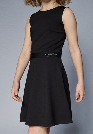 Dziecko w czarnej sukience bez rękawów w kształcie litery A z paskiem z logo Calvin Klein, stojące na jednolitym szarym tle.
