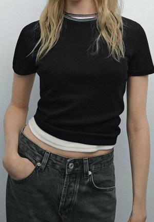 Frau trägt ein schwarzes, geripptes Kurzarmoberteil mit gestreiftem Kragen und hoch taillierte, ausgebleichte schwarze Jeans, Hand in der vorderen Tasche, heller Hintergrund.