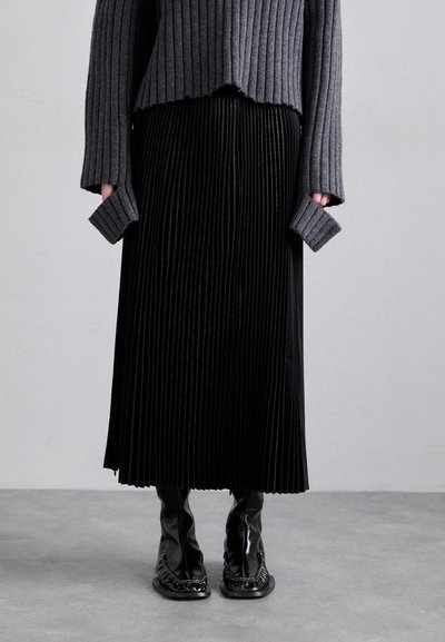 Pull en maille gris à ourlet effiloché associé à une jupe plissée noire. Bottines noires brillantes avec un détail de bordure complexe.