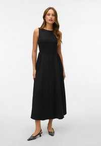 Robe midi sans manches noire avec un buste ajusté et une jupe plissée, fabriquée en tissu lisse. Associée à des chaussures pointues noir et blanc.
