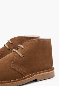 Stivali ankle in suede marrone con due occhielli, lacci beige e dettagli cuciti sulla suola. Il design presenta una punta rotonda e uno stile minimalista.