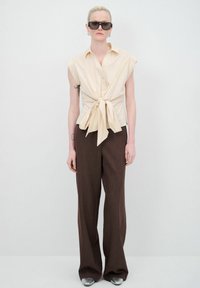 Blusa senza maniche di un beige chiaro con dettaglio nodo frontale, abbinata a pantaloni larghi marroni e scarpe argentate. Design semplice e pulito.