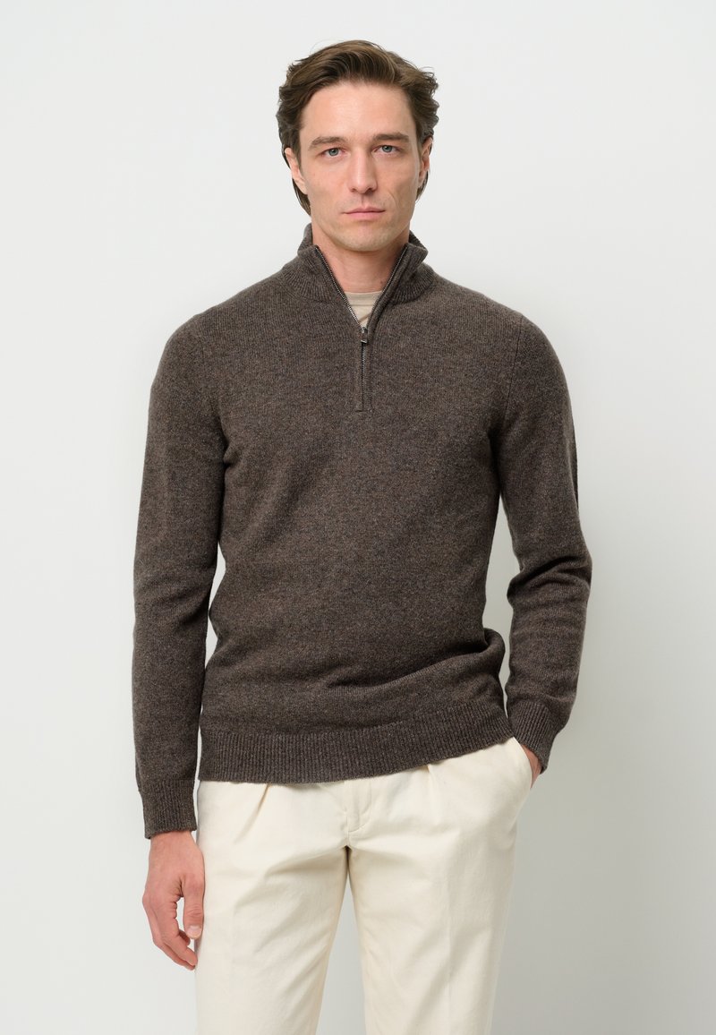 Bruine trui met ritssluiting en geribbelde zoom en manchetten, gemaakt van zachte stof. Gestyled over een lichtgekleurde shirt, gecombineerd met crèmekleurige broek.