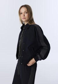 Marine bomberjack met grijze geribbelde accenten, drukknoopsluiting en zijzakken. Gladde textuur en ontspannen pasvorm, gecombineerd met een donker overhemd met knopen.