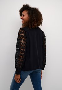 Black shirt featuring intricate lace sleeves with floral patterns, smooth texture, and button closure at the cuff. Worn with blue jeans.