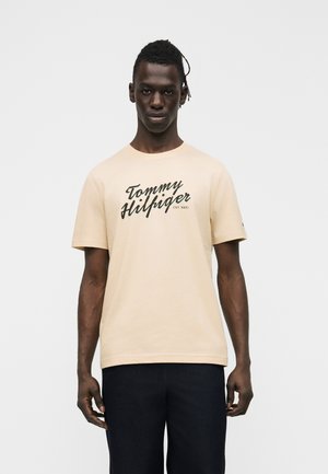 Mann mit dunkler Hautfarbe, der ein beigefarbenes Tommy Hilfiger T-Shirt mit schwarzem Schriftlogo und dunkle Hose trägt, steht vor einem schlichten weißen Hintergrund.