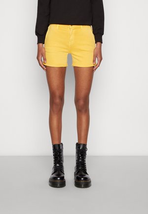 Persona che indossa pantaloncini di jeans gialli, maglia nera a maniche lunghe e stivali neri da combattimento con lacci, in piedi su pavimento grigio contro parete bianca.