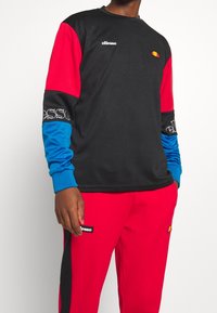 Person som bär en långärmad atletisk tröja i svart, röd och blå samt röda träningsbyxor med synliga Ellesse-logotyper på bröstet, ärmarna och byxorna.