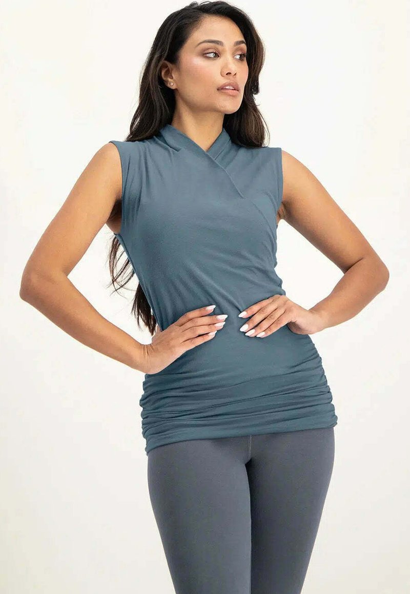 Figurbetontes ärmelloses Oberteil in einem gedeckten Türkis, aus weichem Stoff, mit einem Wickeldesign und Raffung an der Taille. Kombiniert mit grauen Leggings.
