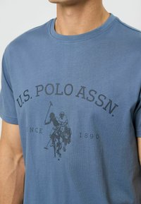 Mėlynas medvilnės marškinėliai su apvaliu kaklu ir atspaudu iš polo žaidėjų grafikos, su užrašu "U.S. POLO ASSN." ir "SINCE 1890".
