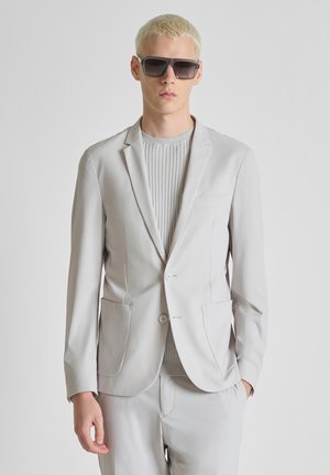 Joven con cabello corto y rubio que lleva grandes gafas de sol oscuras, blazer gris claro, suéter de canalé y pantalones a juego, sobre un fondo liso.