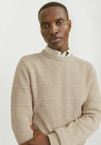 Pull en tricot beige avec des motifs de rayures texturés. Doté d'un col rond et de poignets côtelés. Porté par-dessus une chemise à col rayée.