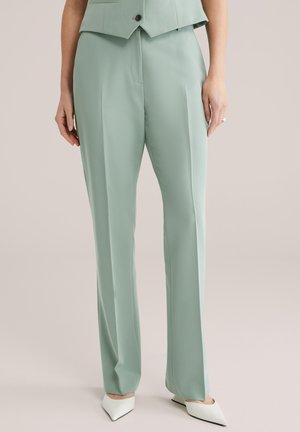Zelené mintové nohavice na mieru z hladkej látky, s voľným strihom, s detailom záhybu a v kombinácii s bielymi lodičkami so špicatou špičkou.