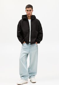 Schwarze, leichte Windjacke mit Kapuze, ausgestattet mit einem vorderen Reißverschluss und Seitentaschen, kombiniert mit hellblauen, locker sitzenden Jeans und weißen Sneakers.