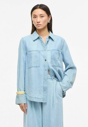 Nő világoskék, túlméretezett farmeringgel, nagy első zsebekkel és hozzáillő bőszárú nadrággal, egyik kézzel zsebében pózol.