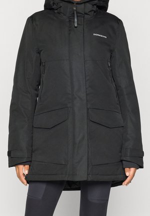 Chaqueta de invierno negra con capucha, cremallera frontal, bolsillos con solapa y correas ajustables en las muñecas, usada por una persona sobre un fondo liso.