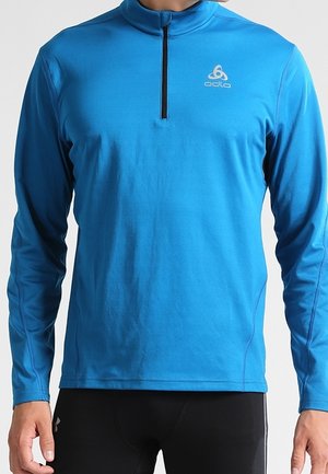 Homme portant un pull-over de sport bleu vif à manches longues avec une fermeture éclair noire sur le haut et un logo blanc sur le côté gauche de la poitrine.