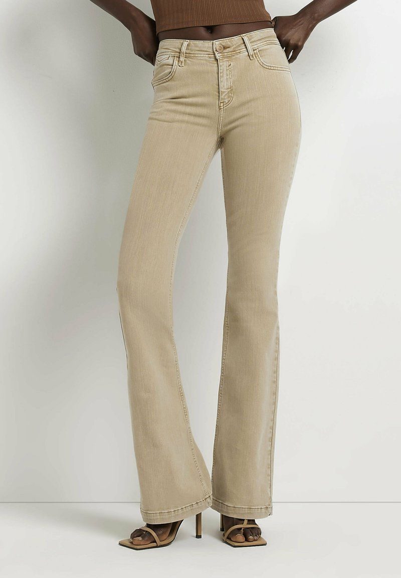 Jean beige slim-fit avec poches avant porté par une personne debout, associé à des sandales à talons hauts à lanières et un top court marron.