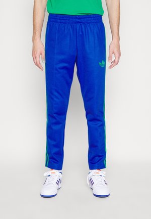 Osoba nosząca niebieskie spodnie dresowe Adidas z zielonymi paskami i białe buty sportowe Adidas, stojąca na jednolitym jasnym tle.