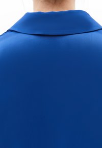 Blusa blu con una texture liscia e lucida, caratterizzata da un colletto classico. Il tessuto appare leggero e morbido, senza motivi o decorazioni visibili.