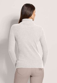 Frau mit langen braunen Haaren, die einen hellgrauen, gerippten Rollkragenpullover und beige Hosen trägt, mit dem Rücken zu einer schlichten Hintergrund.