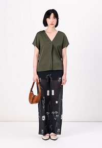 Blusa verde oliva a maniche corte con scollo a V, abbinata a pantaloni neri trasparenti con stampe floreali bianche e una borsa marrone.