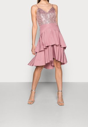 Vrouw draagt een mouwloze roze jurk met een lijfje van pailletten, een asymmetrische gelaagde rok en zilveren hoge hakken met enkelbandje.