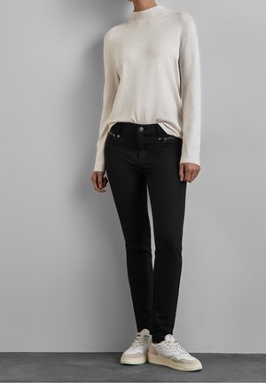 Femme portant un pull à col roulé crème, un pantalon noir slim avec des poches zippées et des baskets blanches, debout contre un mur gris.