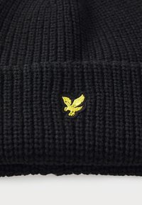 Črna pletena kapa iz teksturiranega materiala, z rumenim vezenim logotipom orla na sprednji strani in z zavihkom.