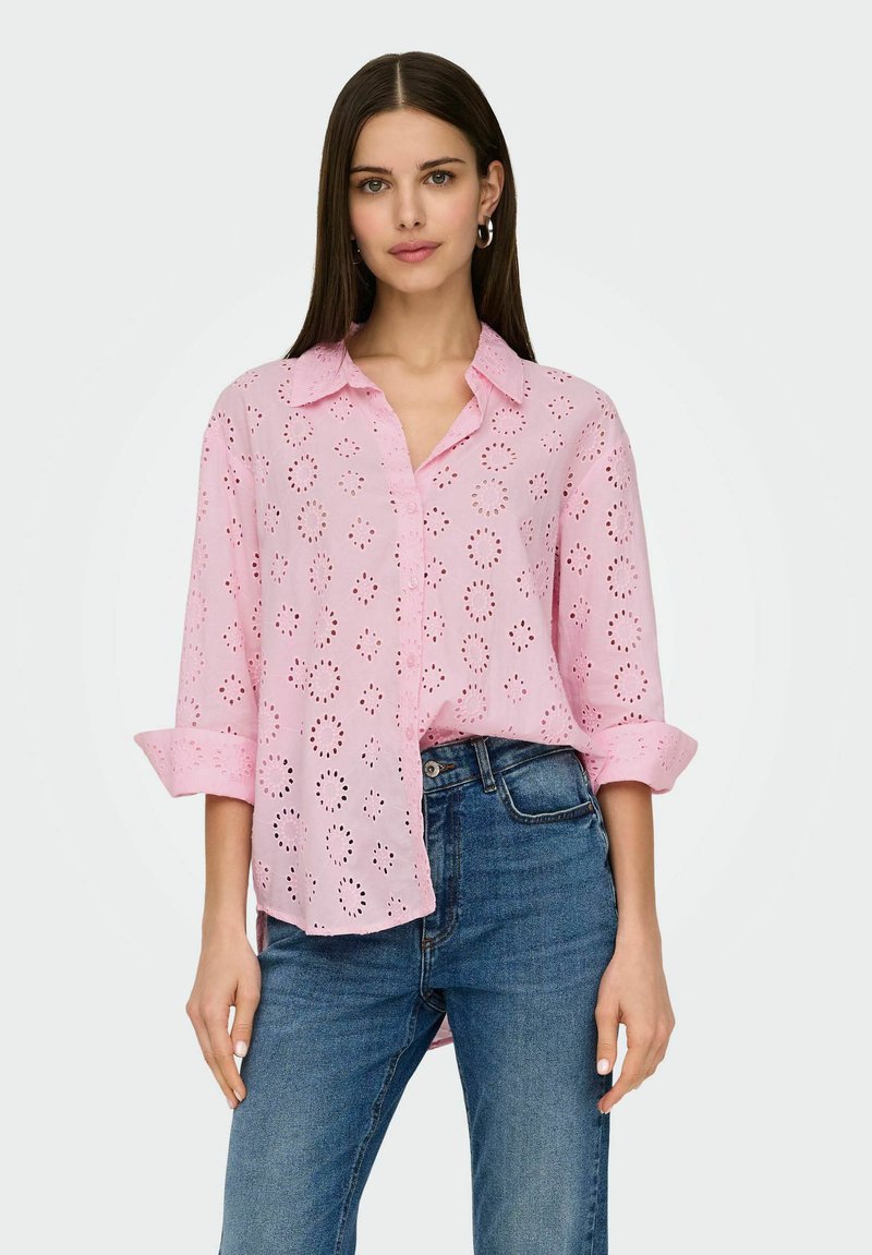 Camicia rosa chiaro con bottoni e motivo ad occhio, con maniche arrotolate. Abbinata a jeans di denim blu, che mostrano una vestibilità rilassata e uno stile casual.