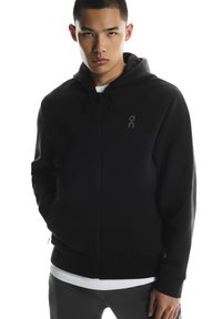 Musta vetoketjullinen huppari, joka on valmistettu pehmeästä kankaasta. Siinä on kengurun tasku, säädettävä huppu nyörillä ja hillitty logo rinnassa. Tasasaumat näkyvissä.