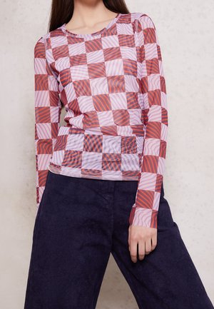 Femme portant un haut à manches longues rose et rouge à carreaux avec des motifs abstraits, assorti à un pantalon large foncé, debout devant un mur pâle.